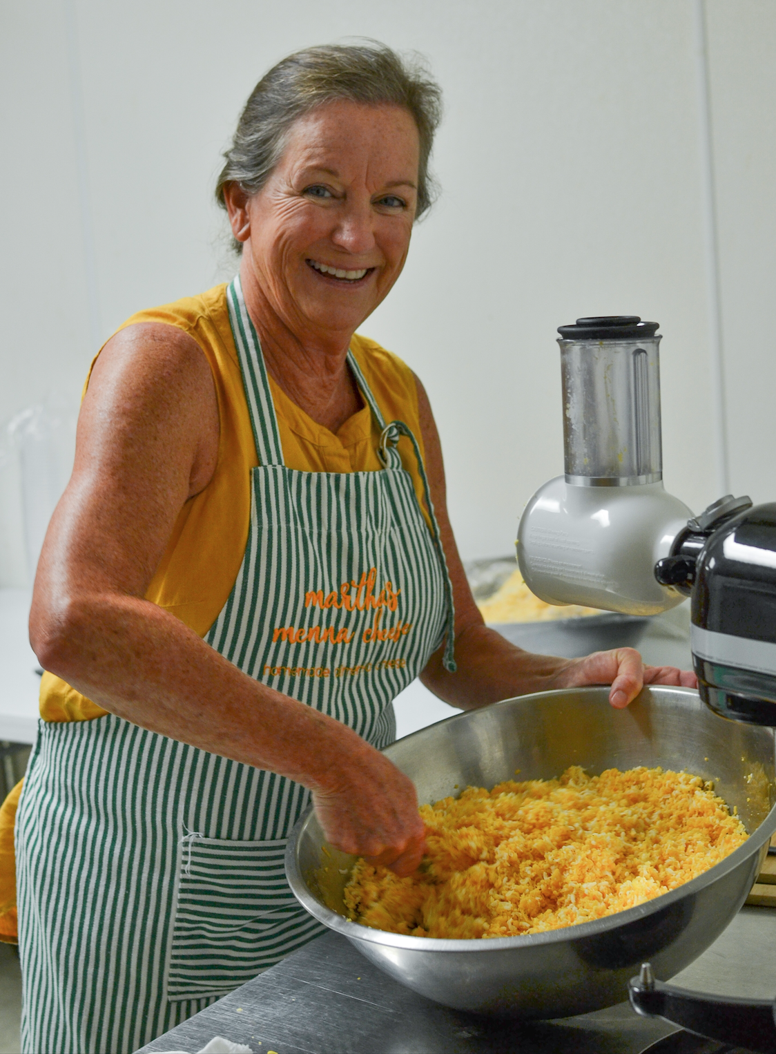 Smiling woman in a striped apron mixing ingredients in a large bowl, showcasing a kitchen setting with a food processor and a stainless steel counter, emphasizing cooking and food preparation.