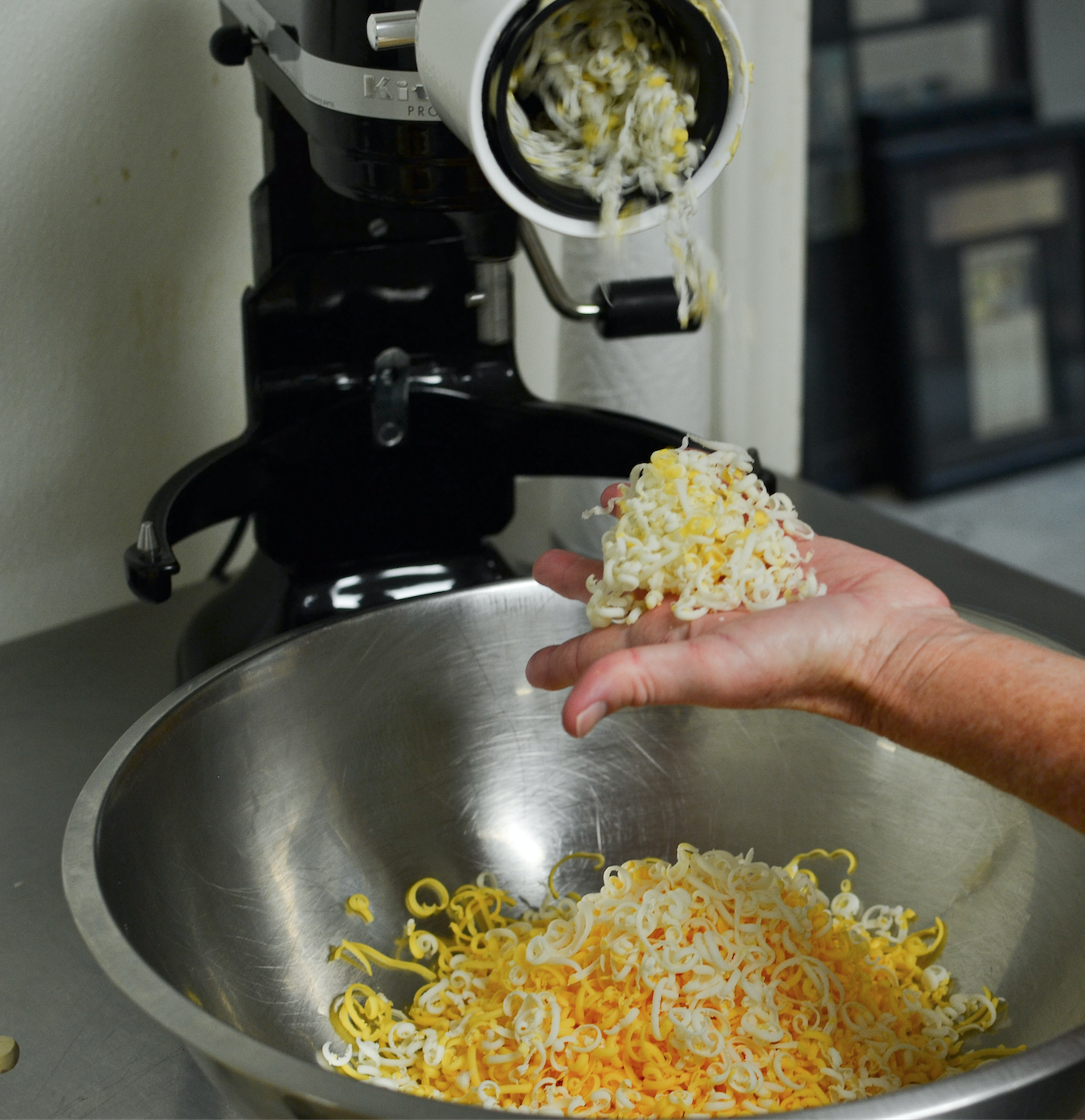 Hand holding freshly grated cheese while a mixer dispenses more cheese into a large metal bowl, showcasing the process of preparing ingredients for cooking.